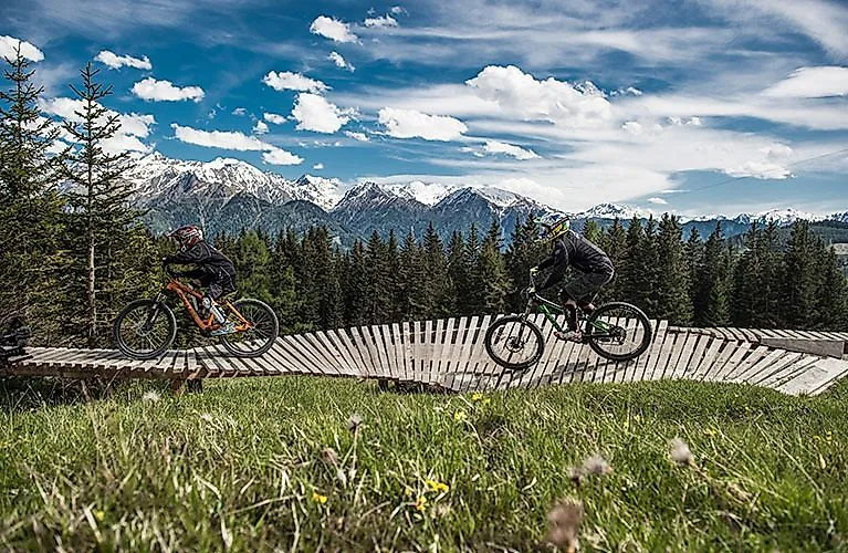 Zwei Mountainbiker fahren auf einem Holztrail im Bikepark Serfaus-Fiss-Ladis mit Blick auf die verschneiten Alpen.