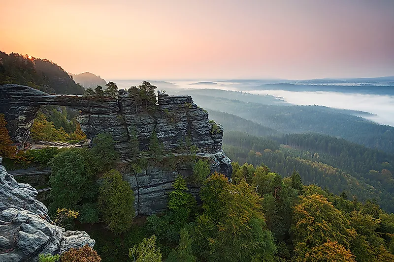 Felsformation Basteibrücke im Elbsandsteingebirge bei Sonnenaufgang mit Blick über bewaldetes Tal im Nebel