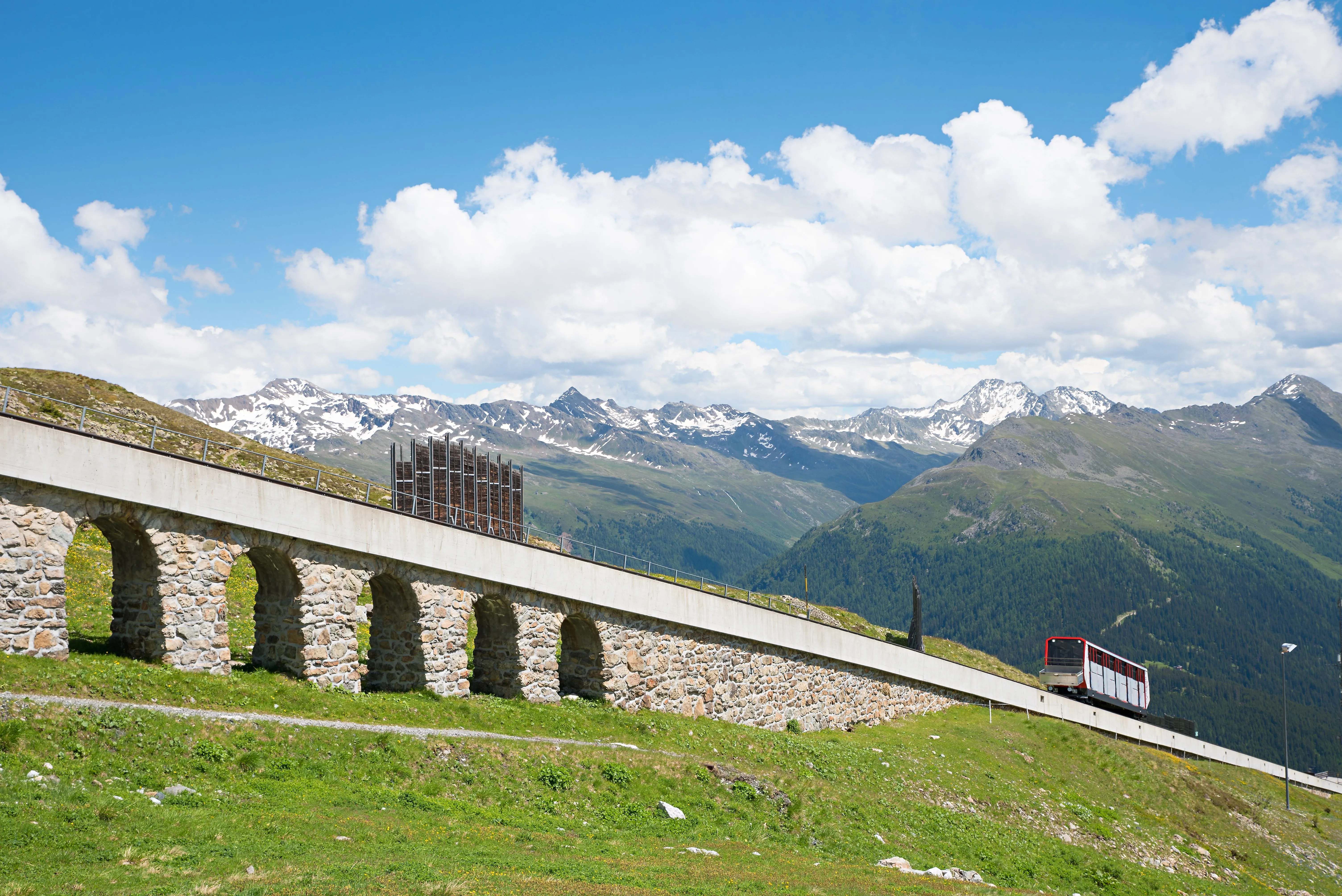 Die Standseilbahn Parsennbahn in Davos im Sommer, umgeben von grünen Almwiesen und den schneebedeckten Gipfeln der Alpen.