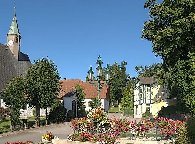 Place de village estivale à Mönichkirchen avec des parterres de fleurs, des bâtiments historiques et un ciel bleu.