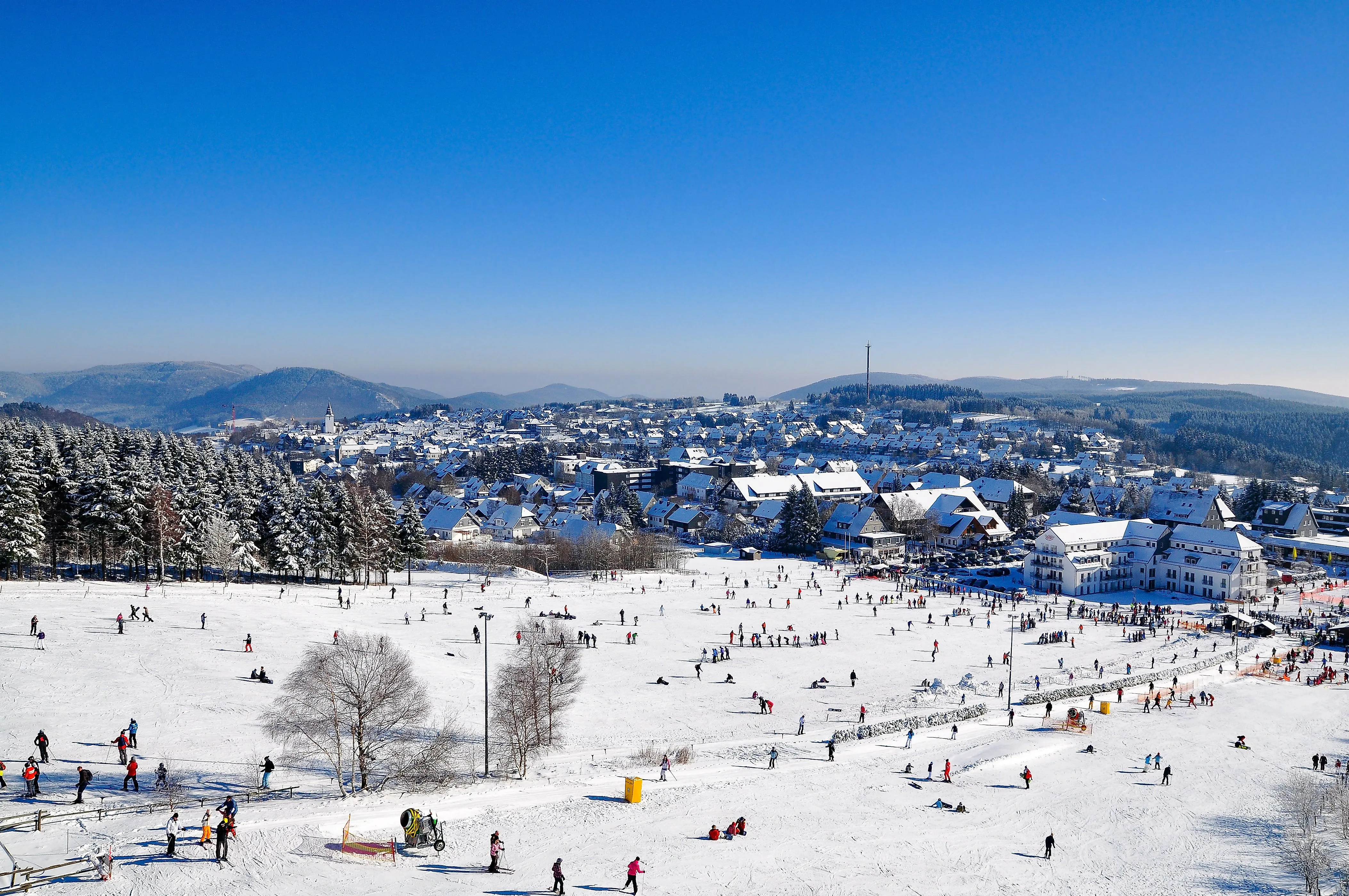 Wintersportgebiet in Winterberg mit verschneiter Piste, Skifahrern und Blick auf die Stadt im Hintergrund.