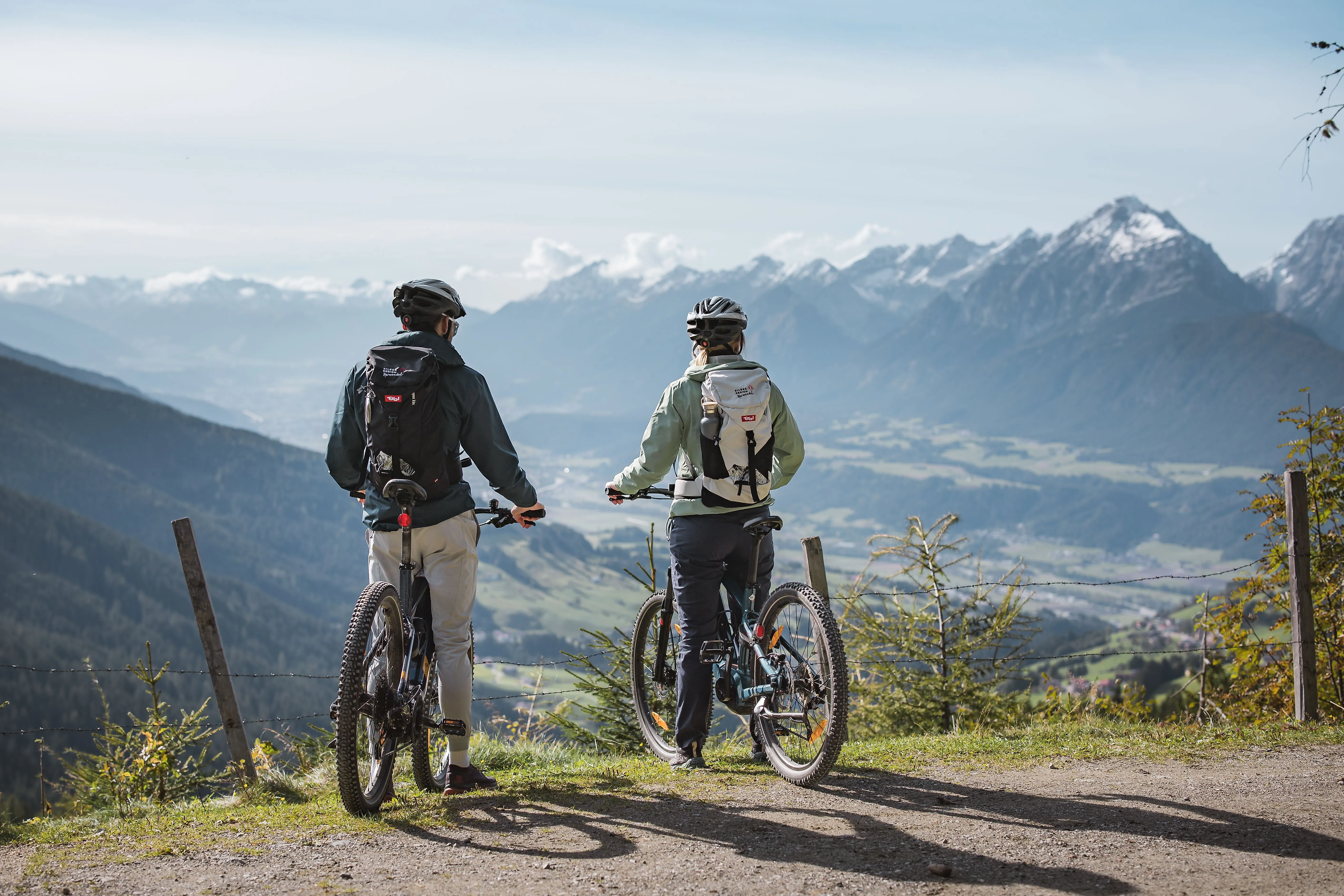 Dos ciclistas de montaña con bicicletas en un mirador con vistas al valle y panorama alpino al fondo