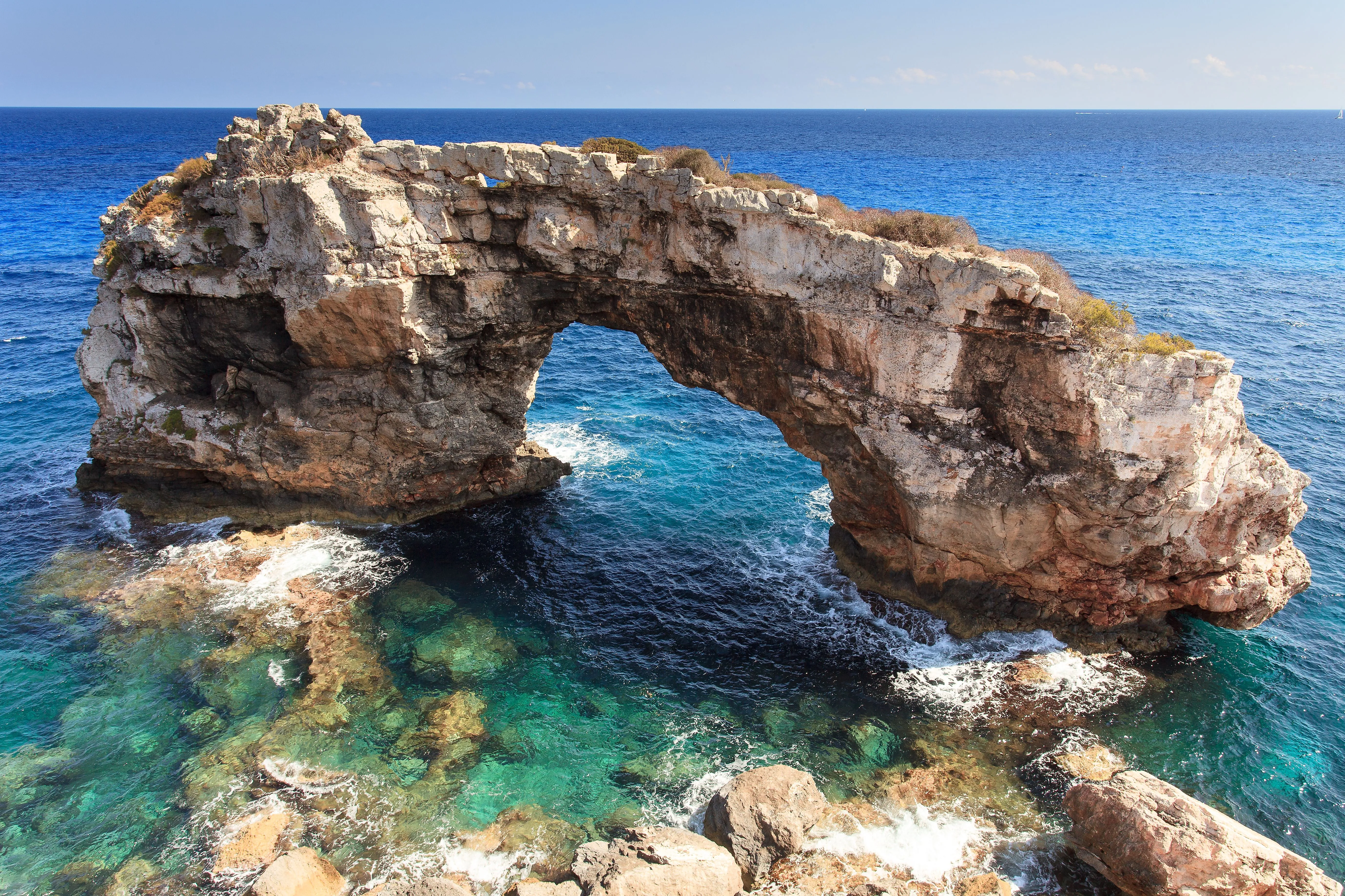 Natural rock bridge Es Pontàs off the coast of Mallorca with crystal clear water and rocks in the foreground.