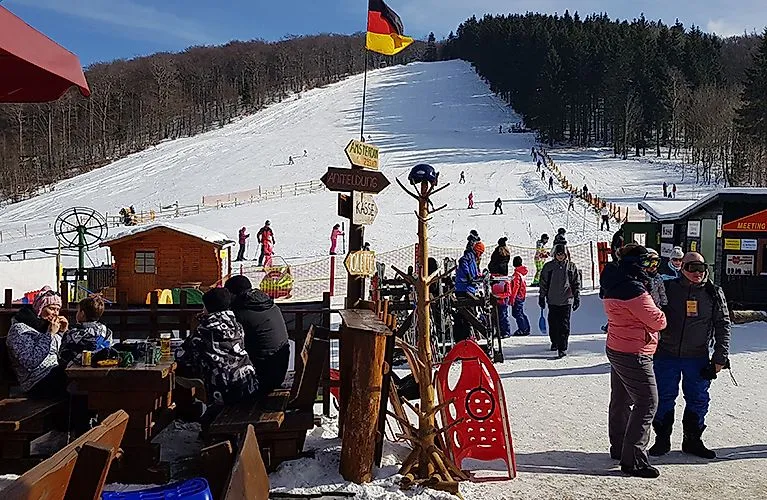 Winterlicher Tag im Familienskigebiet Schlossberg in Medebach mit Skifahrern, Rodlern und Besuchern vor der Hütte. Im Hintergrund der Skihang und ein Sessellift.