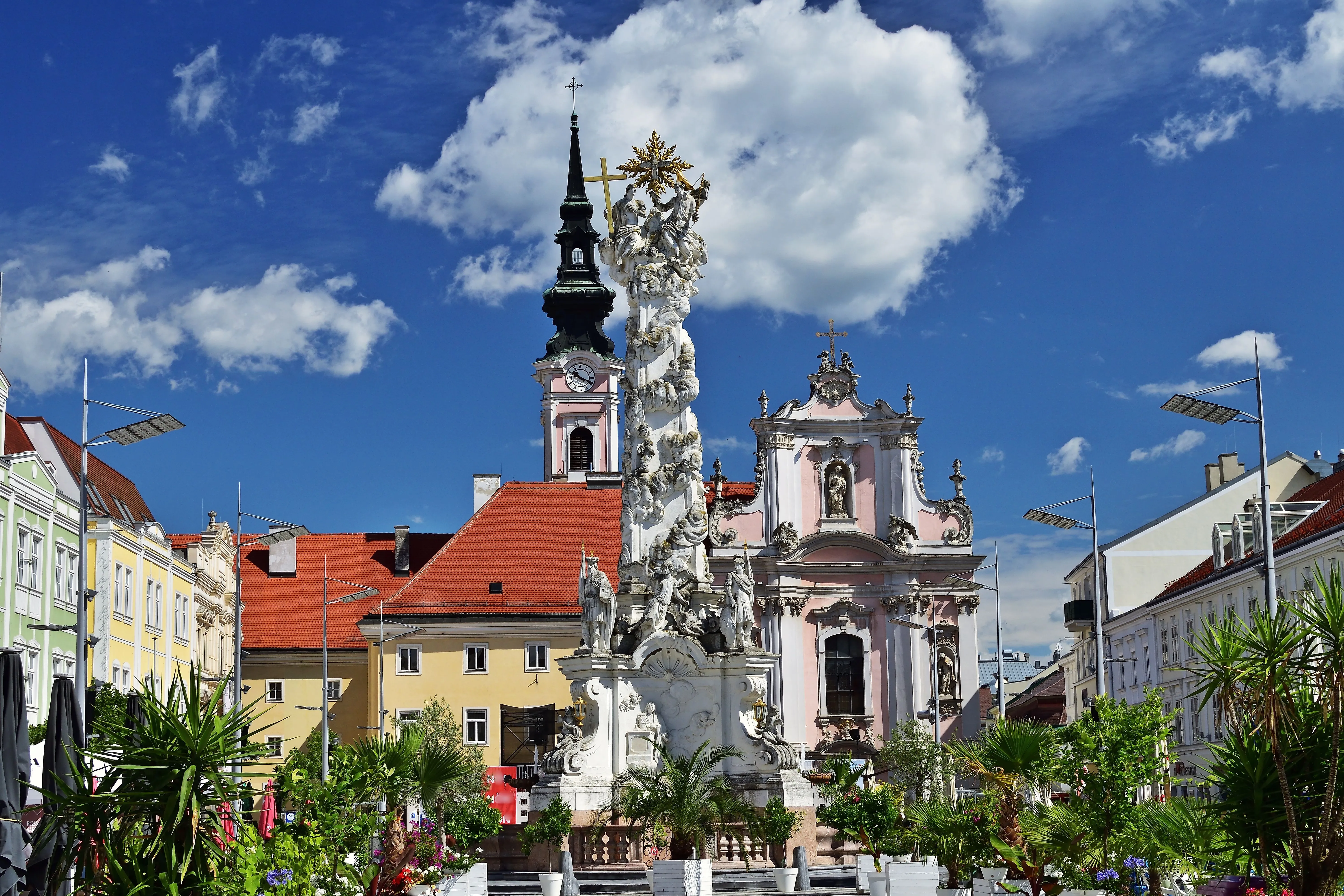 Historický sloup Nejsvětější Trojice před barokním františkánským kostelem v centru St. Pöltenu, obklopený barevnými měšťanskými domy a letní květinovou výzdobou.