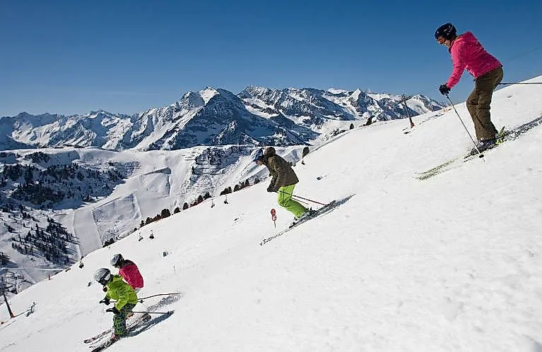 Una familia esquiando junta en una pista soleada con un magnífico panorama alpino de fondo.