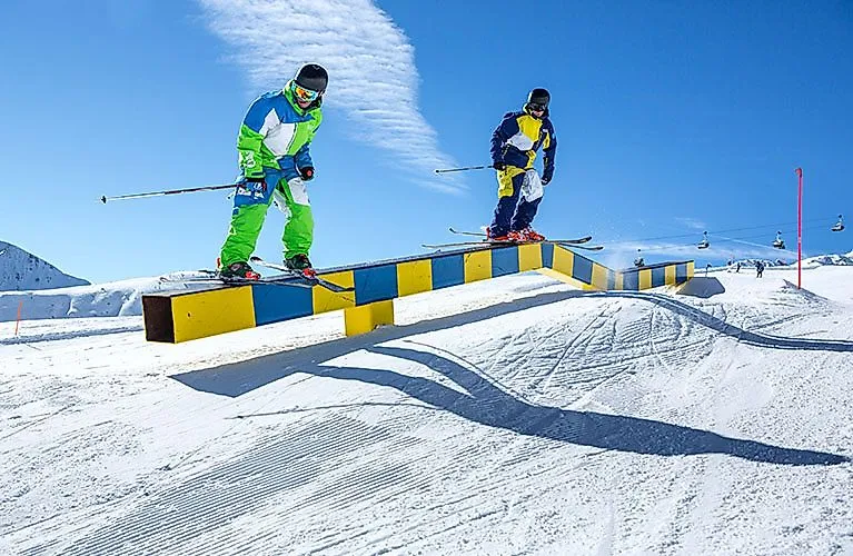 Zwei Skifahrer springen synchron über eine bunte Rail im Snowpark von Serfaus-Fiss-Ladis bei strahlendem Sonnenschein.
