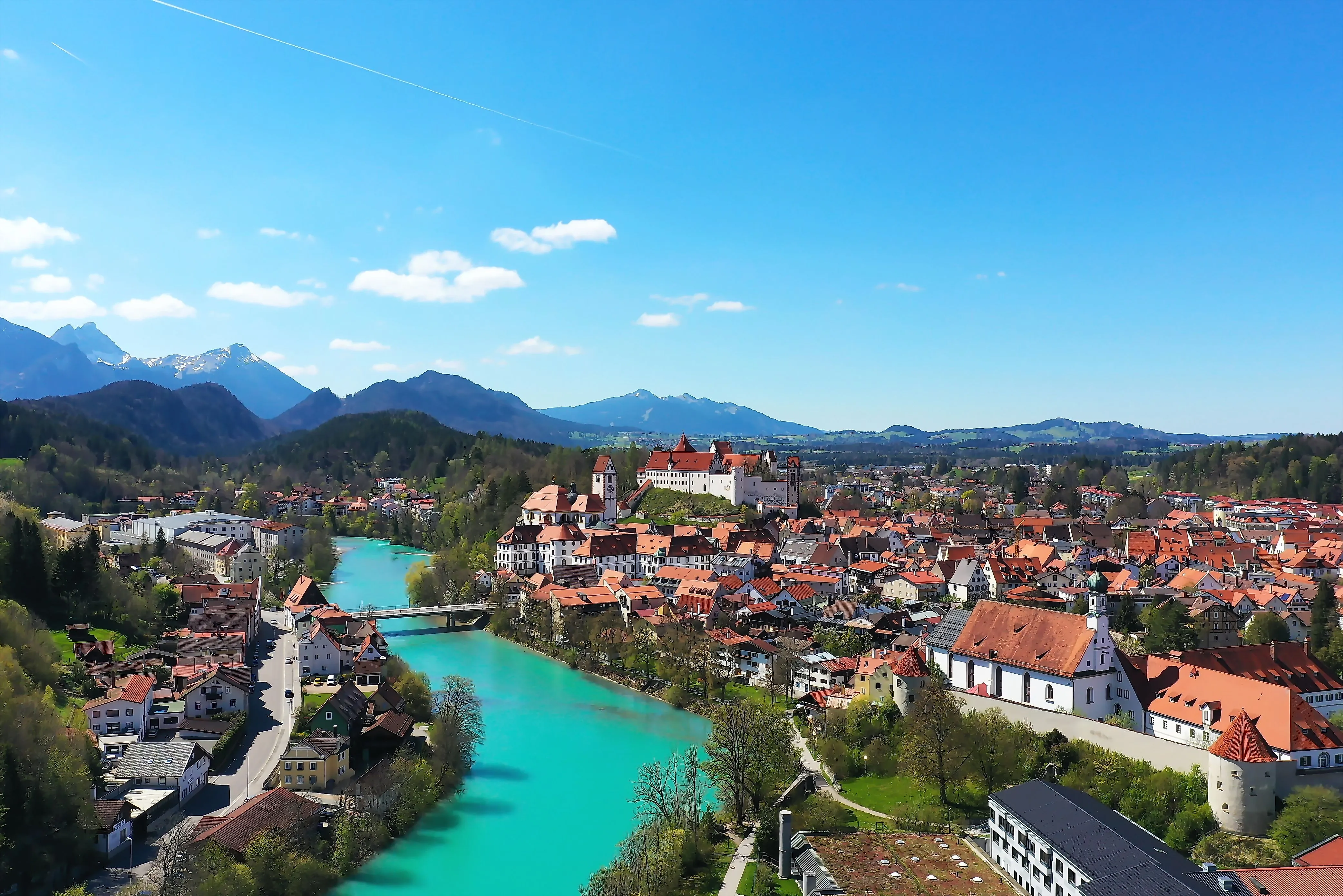 Panoramaaufnahme von Füssen mit dem türkisfarbenen Lech, historischen Gebäuden, dem Hohen Schloss und Alpenkulisse im Hintergrund