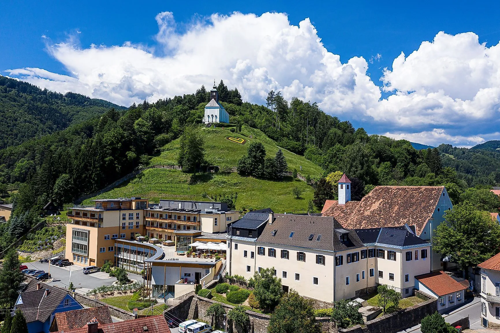 Aerial view of the Heilmoorbad in Bad Schwanberg with church on the Rosenkogel and wooded hilly landscape in the background.