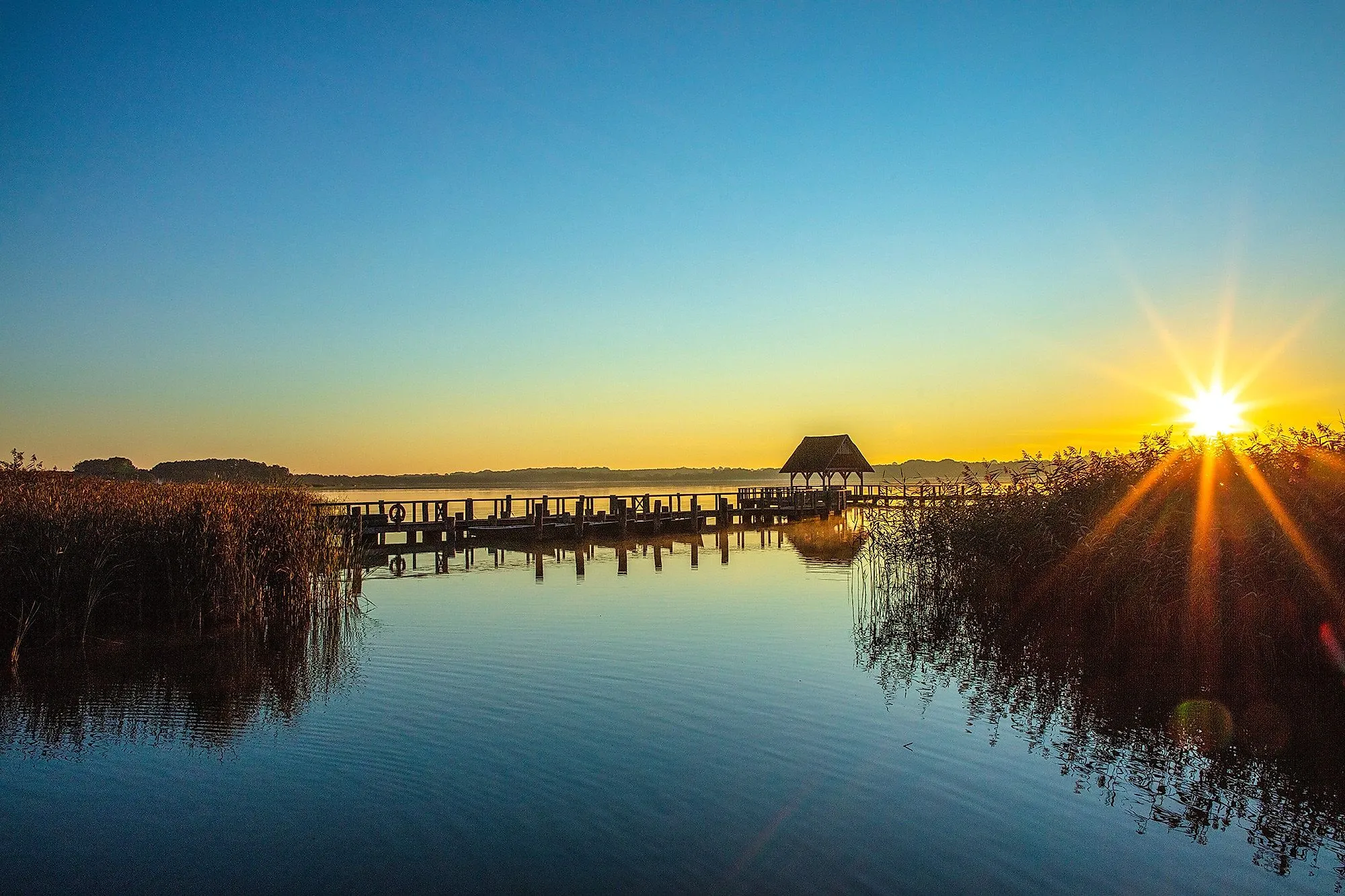 Západ slunce na jezeře Hemmelsdorf s molem a rákosím v teplém večerním světle.