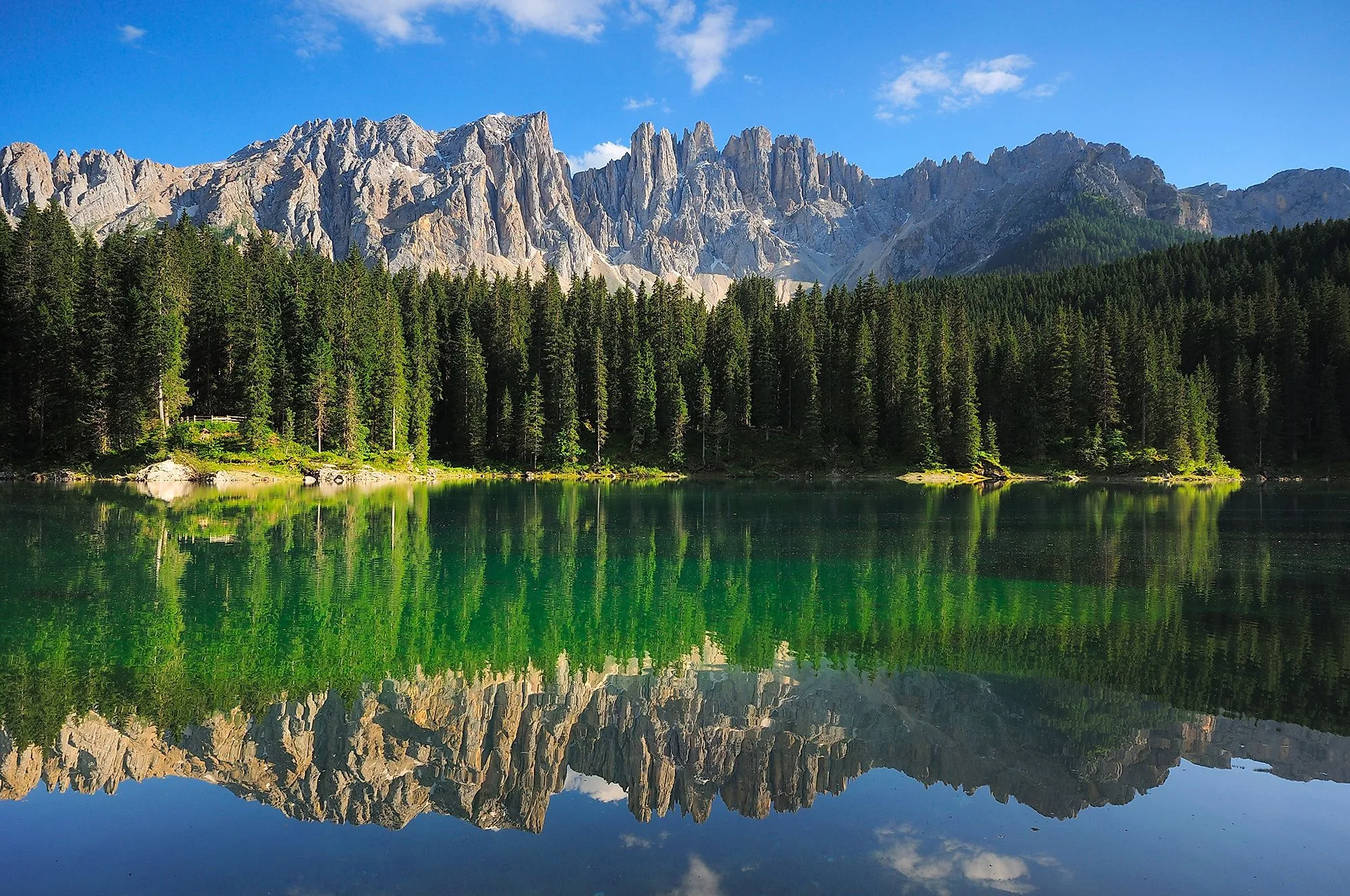 Kristalhelder Carezza meer met een weerspiegeling van de Latemar bergen in Zuid-Tirol, omringd door dichte bossen en blauwe lucht.