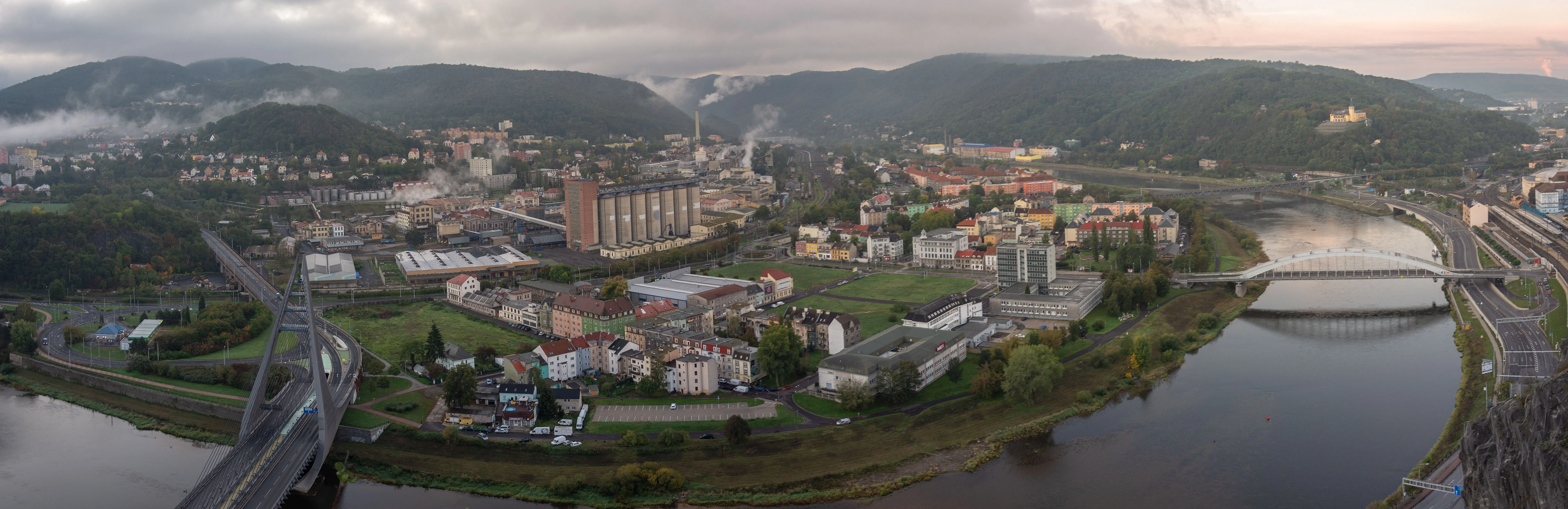 Stadt an der Elbe mit Brücken, Industrieanlagen und Wohnhäusern vor bewaldeten Hügeln bei Nebel