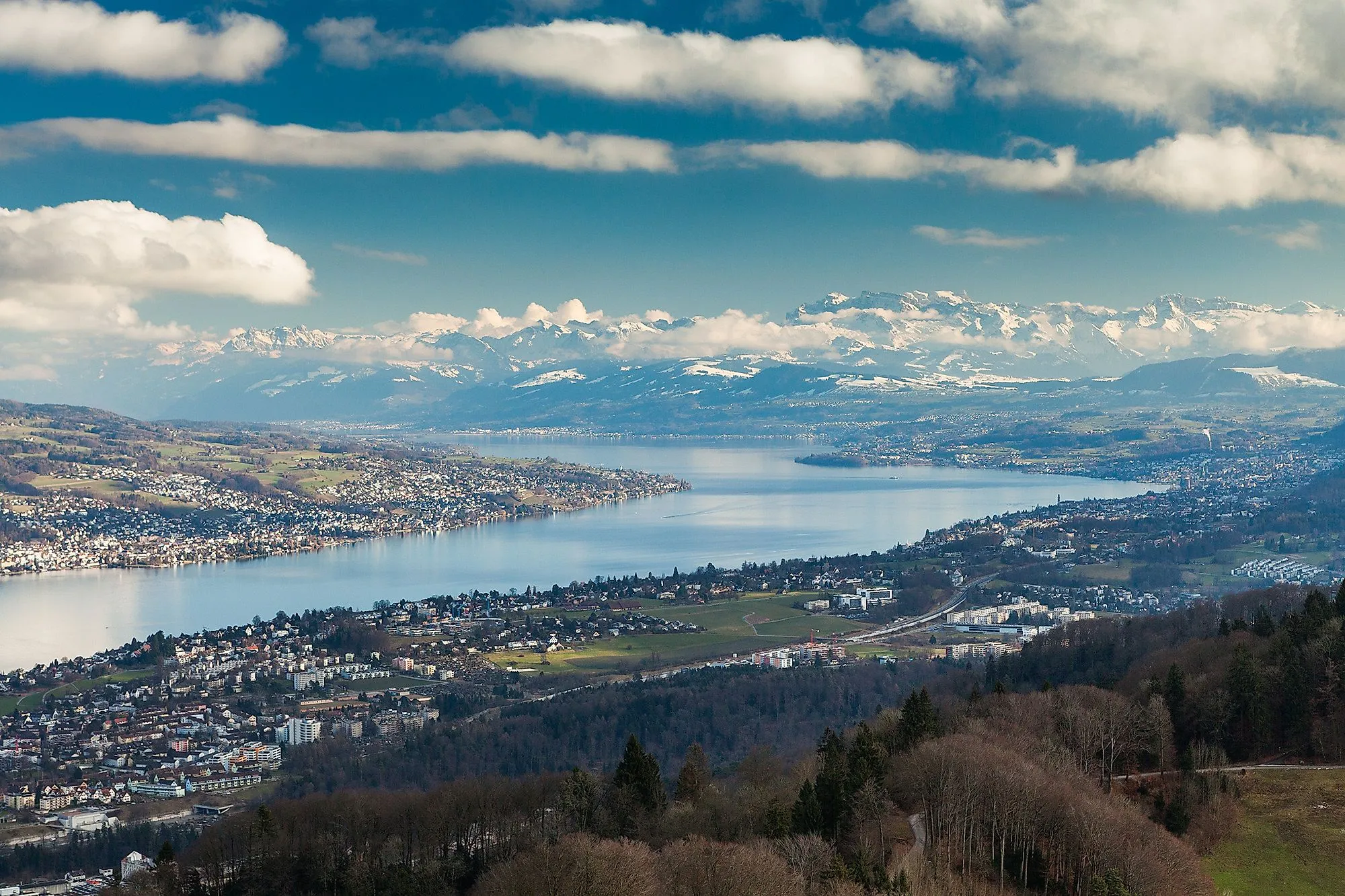 Far-reaching view of Lake Zurich with lakeside villages and the snow-covered Alps in the background on a partly cloudy day.
