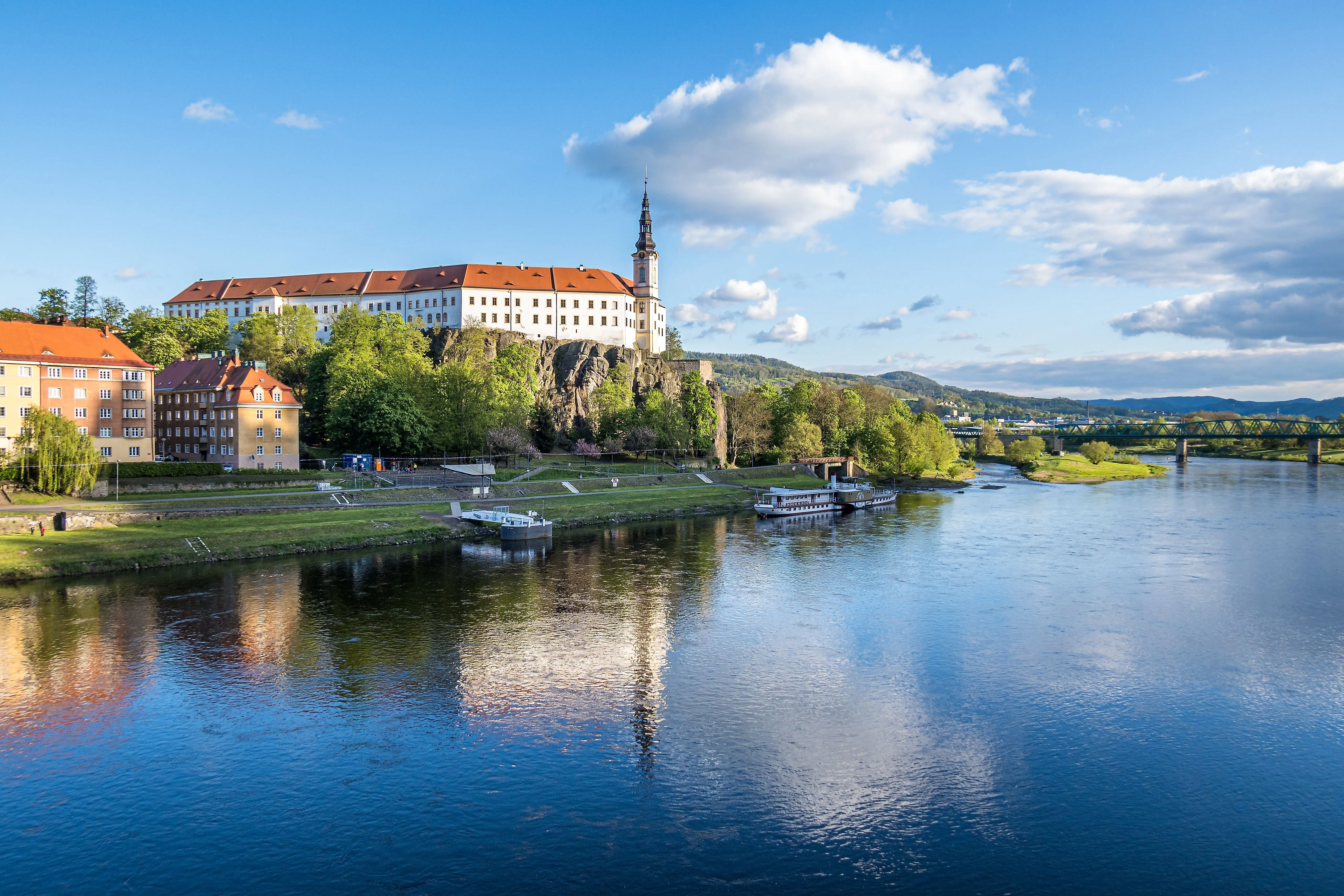 Schloss auf Felsen an der Elbe in Děčín mit Fluss, Boot und Brücke unter blauem Himmel