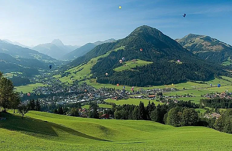 Summer panorama of Kirchberg in Tirol with green alpine meadows, surrounding mountains and numerous hot air balloons in the sky.
