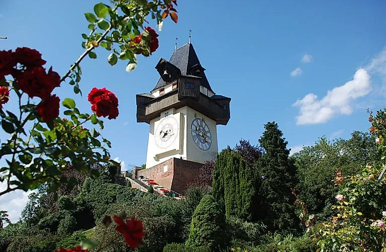 Historická hodinová veža na Schlossbergu v Grazi, obklopená kvitnúcimi ružami a zelenou vegetáciou počas slnečného letného dňa.