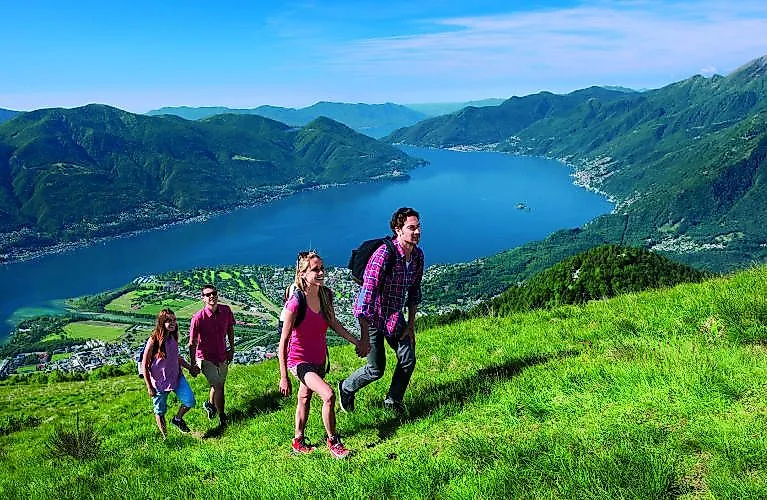 Turistická skupina s rodinou si užíva slnečný letný deň na zelenej alpskej lúke s panoramatickým výhľadom na jazero Maggiore neďaleko Minusio.