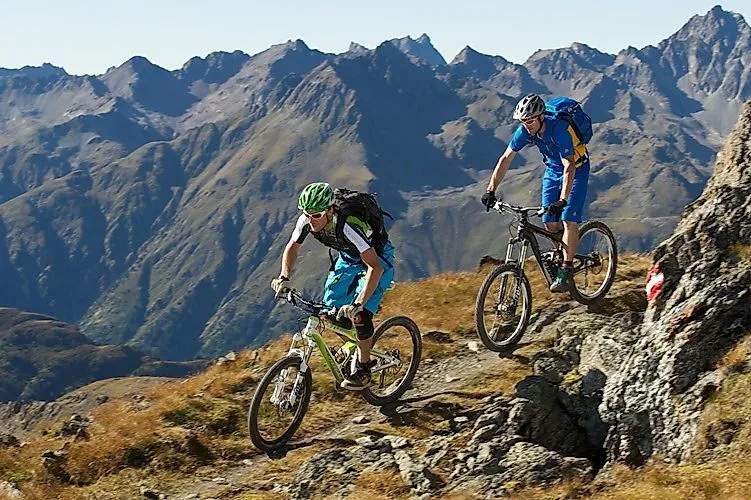 Zwei Mountainbiker fahren auf einem alpinen Trail oberhalb von Ischgl, umgeben von imposanten Bergrücken in der Tiroler Alpenlandschaft.