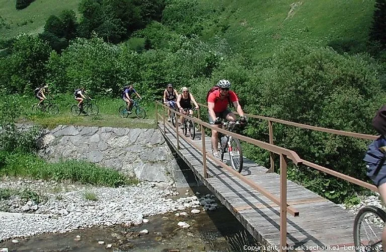 Gruppe von Mountainbikern überquert eine schmale Holzbrücke über einen Gebirgsbach in Berwang, umgeben von grüner Alpenlandschaft im Sommer.