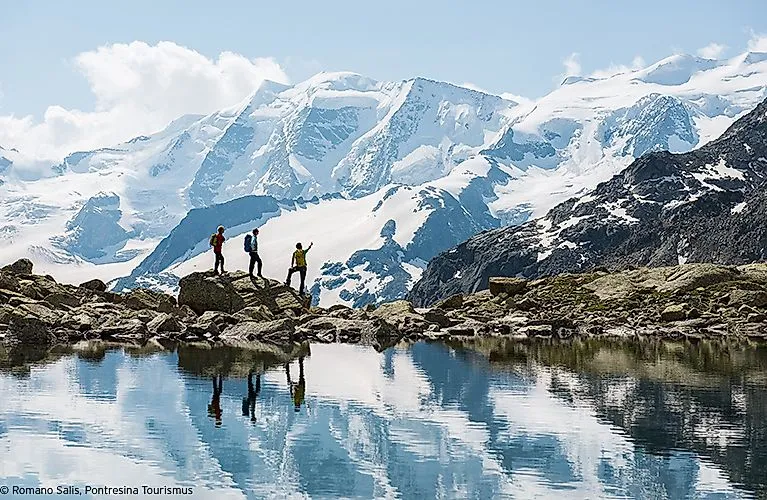 Tři turisté stojí na skalách u jezera v oblasti Pontresina, obklopeni zasněženými horami a odrazem ve vodě.
