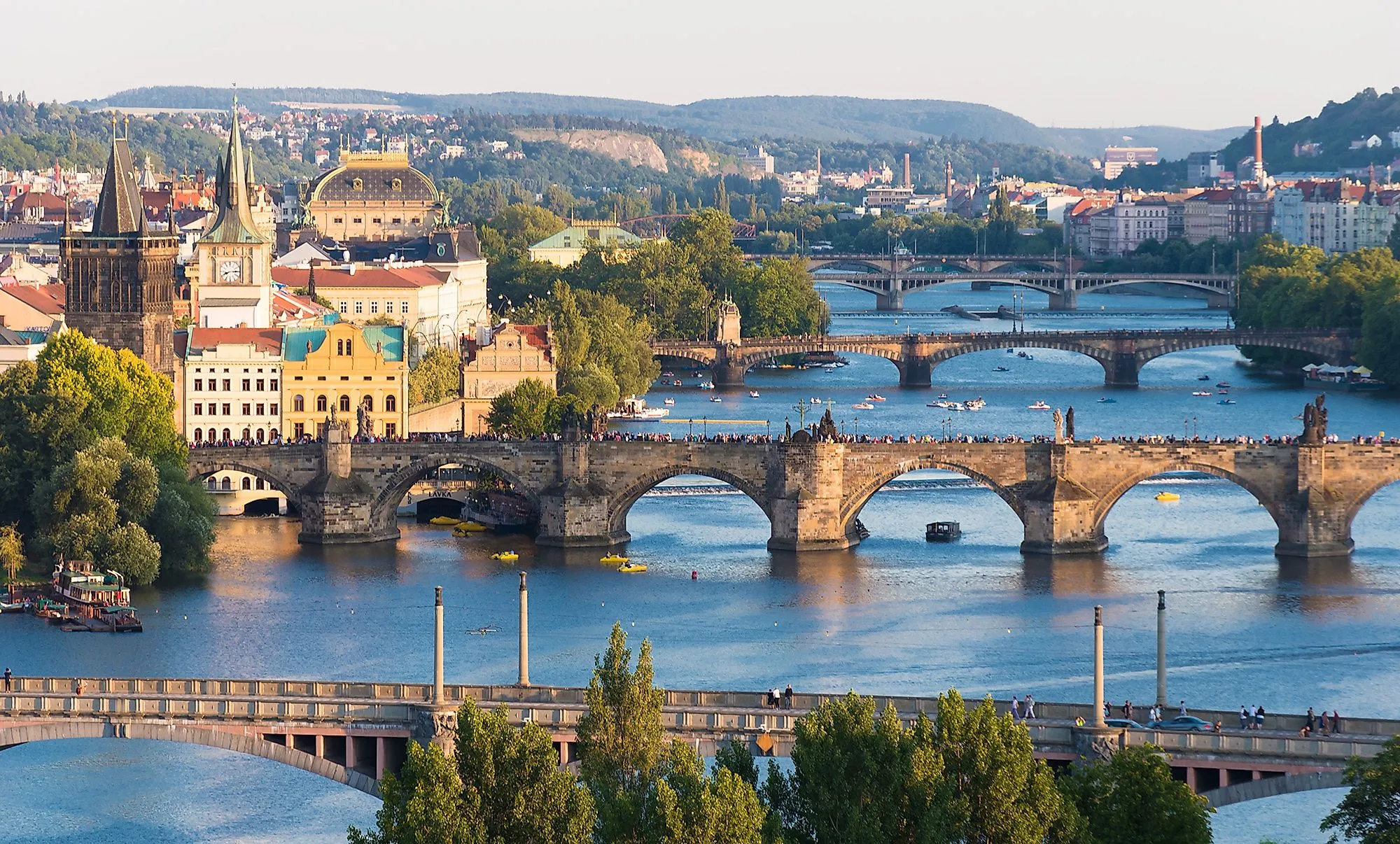 Pohled na Karlův most přes Vltavu v Praze s věžemi a mnoha loděmi.