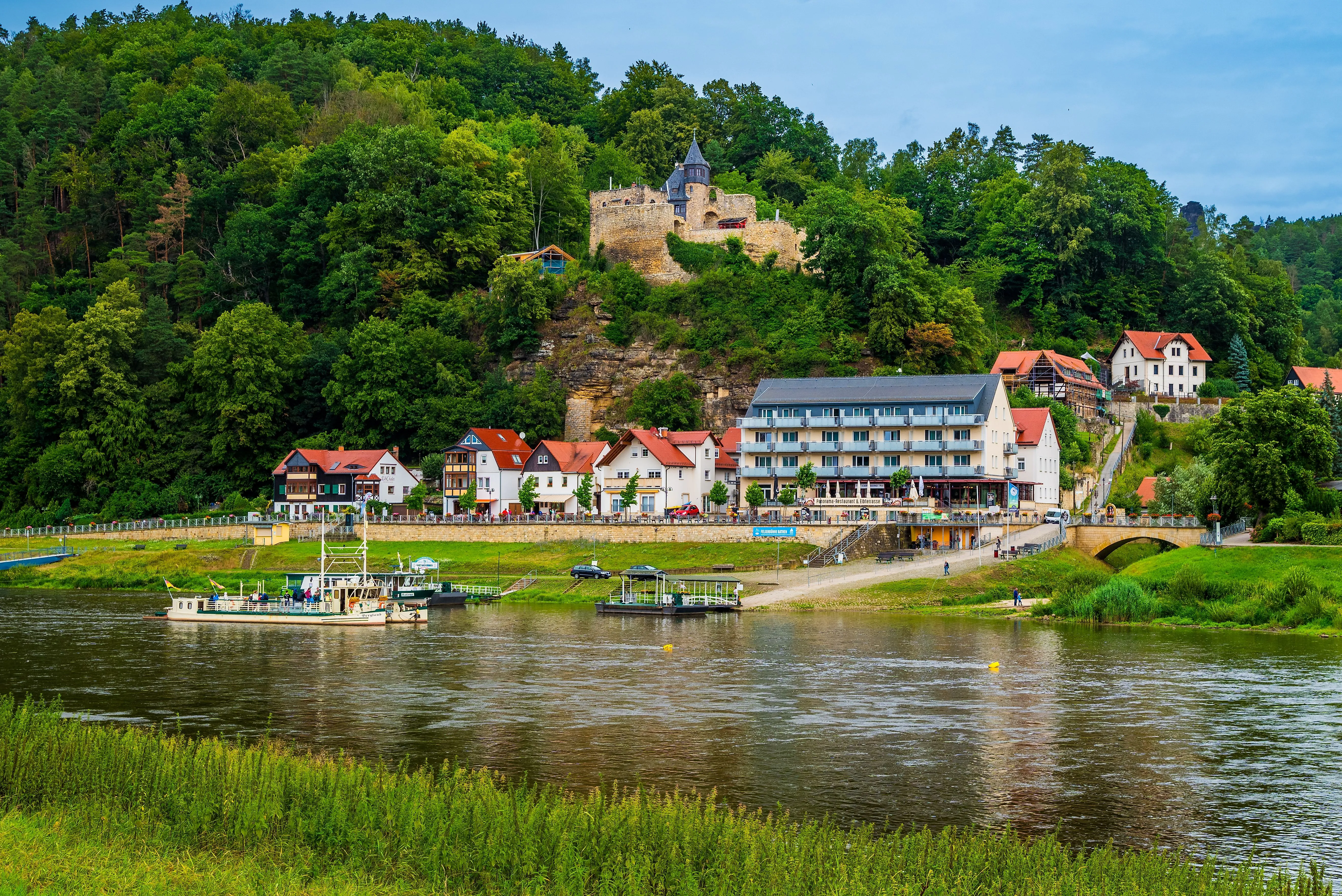 Uitzicht op de idyllische oevers van de Elbe met de veerboot in Rathen, omringd door vakwerkhuizen en de schilderachtige achtergrond van Kasteel Altrathen.