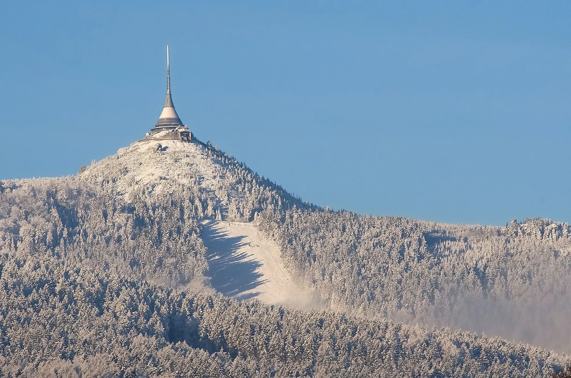 Zasnežená hora Ještěd s televíznou vežou a hotelom na vrchole pod modrou oblohou.