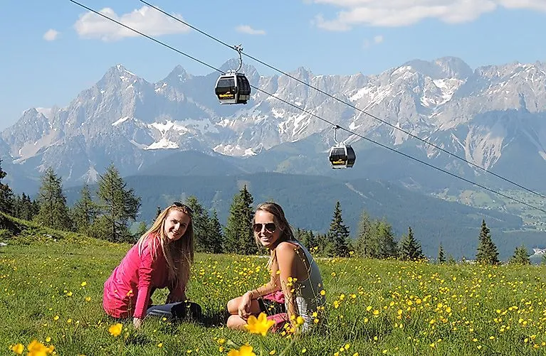 ALT-Text:Two young women enjoy the view on the Reiteralm in summer, with a view of the imposing Dachstein mountains and passing gondolas.