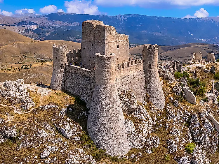Středověký hrad na horském hřebeni v alpské krajině se skalami a širokými kopci pod modrou oblohou