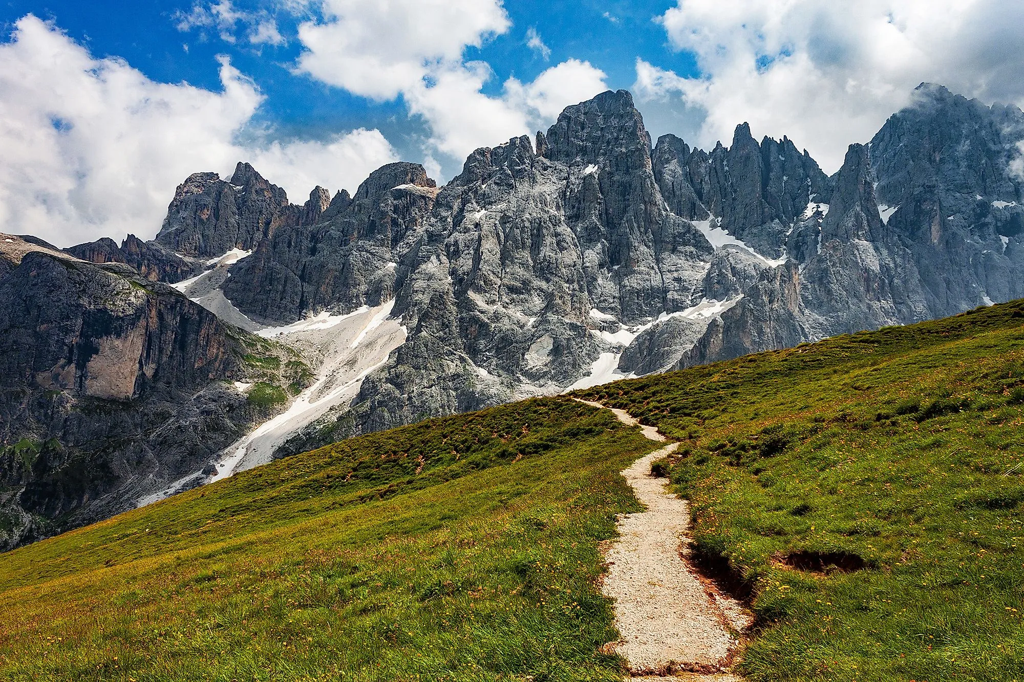 Turistická stezka na zelené horské pastvině před působivou skalní kulisou San Martino di Castrozza v Dolomitech.