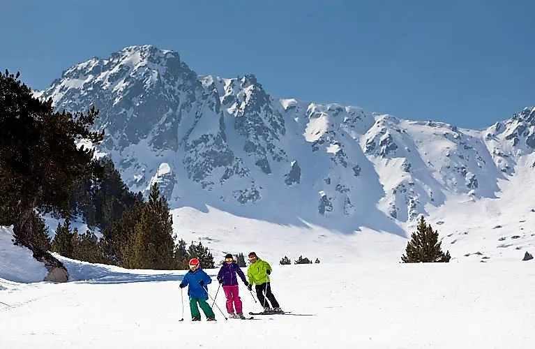 Family skiing in the sunshine on a wide ski slope in Grandvalira with snow-covered mountains in the background.
