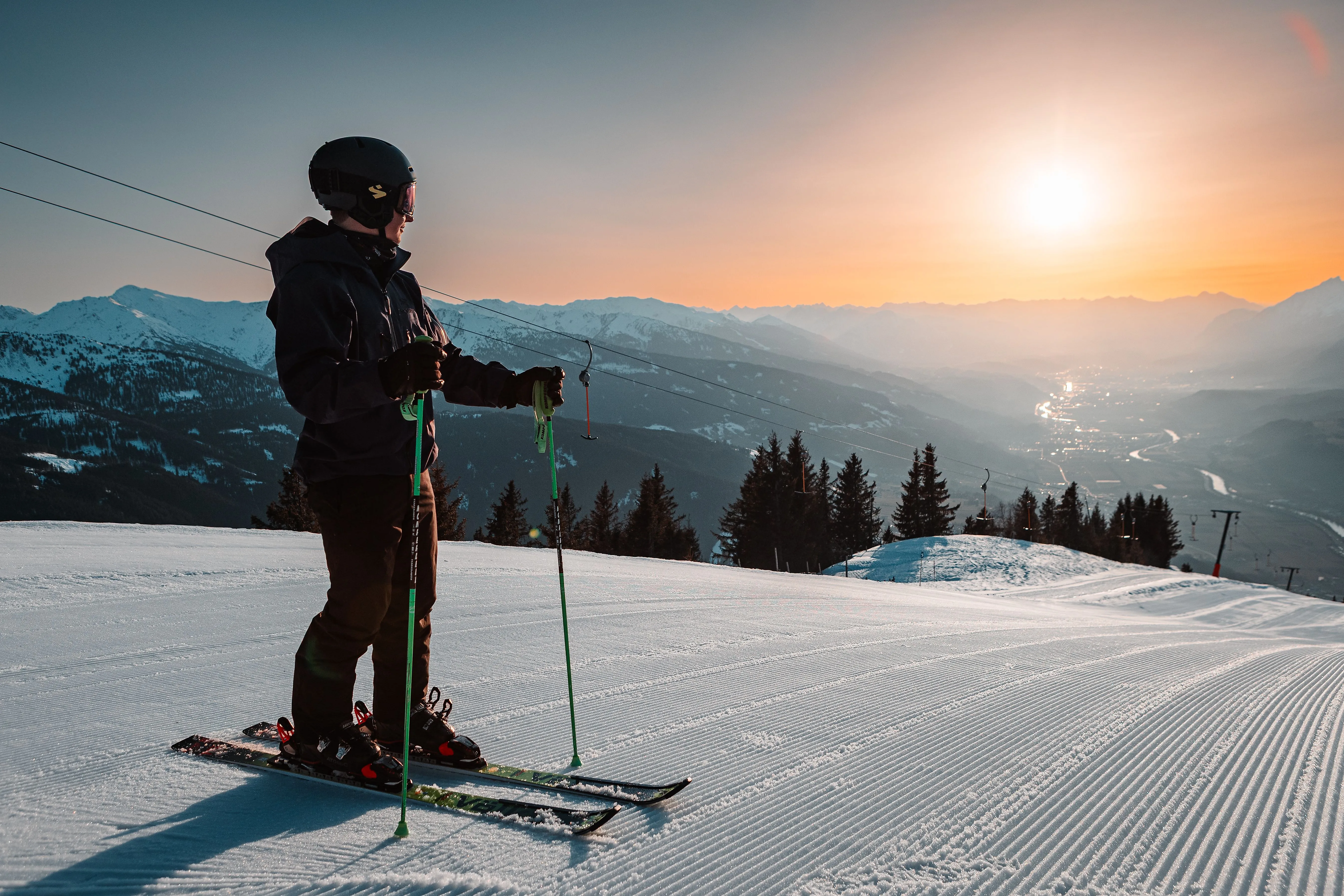 Esquiadores en una pista preparada al atardecer con una vista de los Alpes nevados y el valle al fondo.