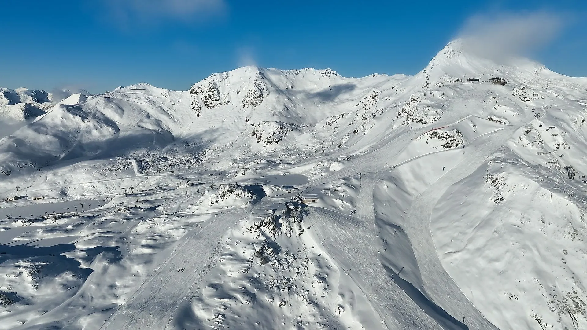 FlyingCam shot of the snow-covered mountain landscape and pistes in the Obertauern ski resort under a bright blue sky.