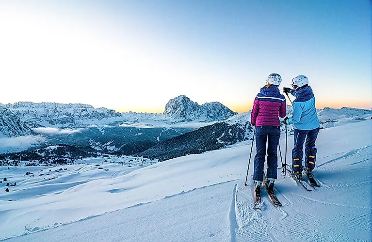 Two skiers on a snow-covered slope with a view of the Dolomites at sunrise