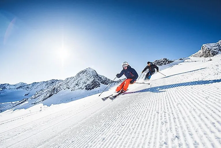 Two skiers on a freshly groomed piste on the Stubai Glacier with bright sunshine and snow-covered mountain peaks.