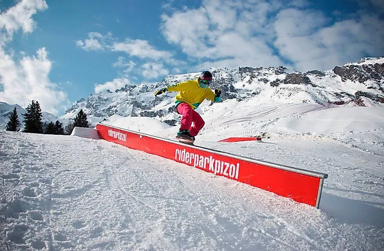 Snowboardisté předvádějí triky v Riderparku Pizol na pozadí zasněžených hor.