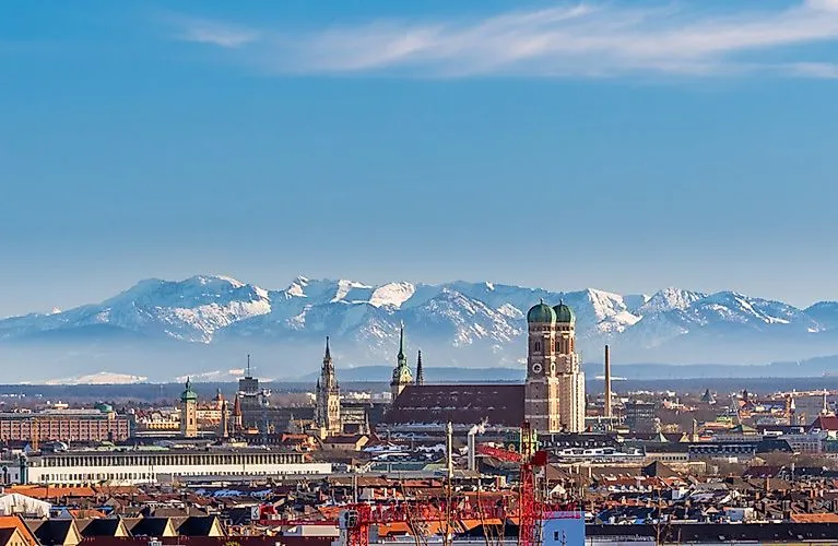 View of the Munich skyline with the Frauenkirche in the foreground and the snow-covered Alps in the background under a clear sky.