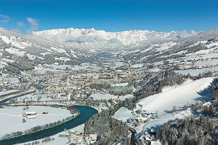 Zasněžená zimní krajina s výhledem na St. Johann im Pongau a okolní hory pod jasnou modrou oblohou.