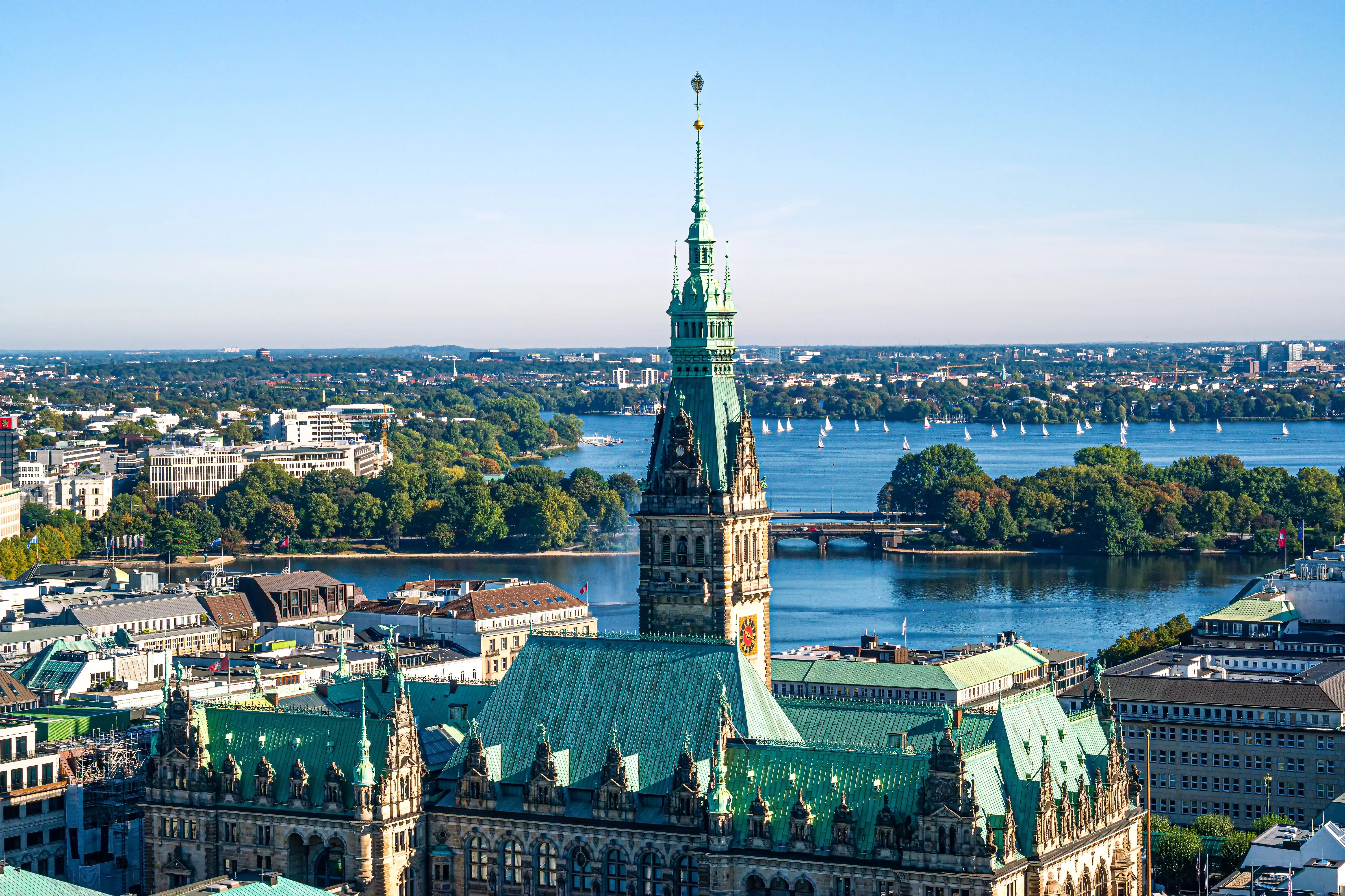 View over Hamburg with the historic town hall in the foreground and the Outer Alster in the background, on which numerous sailing boats are underway.