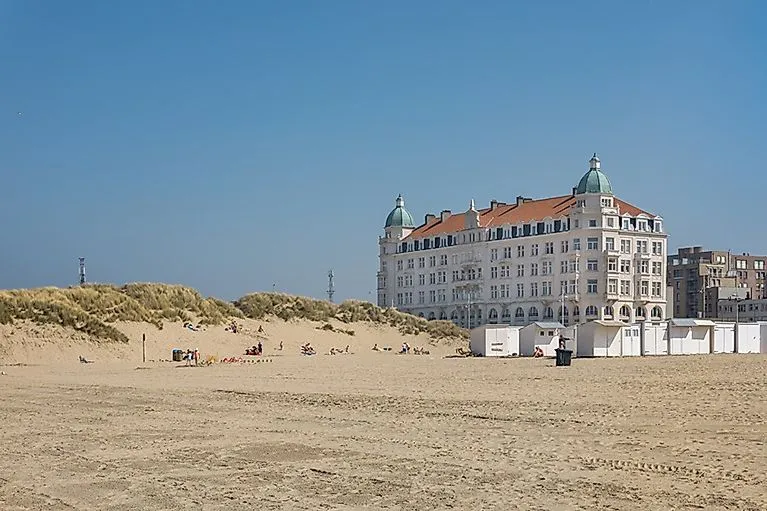 Bright sandy beach panorama of Zeebrugge with some bathers and a historic white hotel building with green domed roofs in the background under a blue sky.