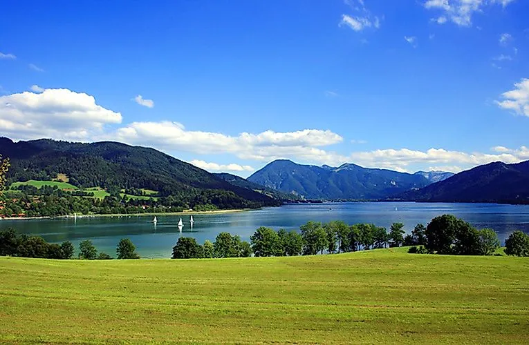 Vast summer landscape with a view of Lake Tegernsee, lined with green meadows and wooded mountains.