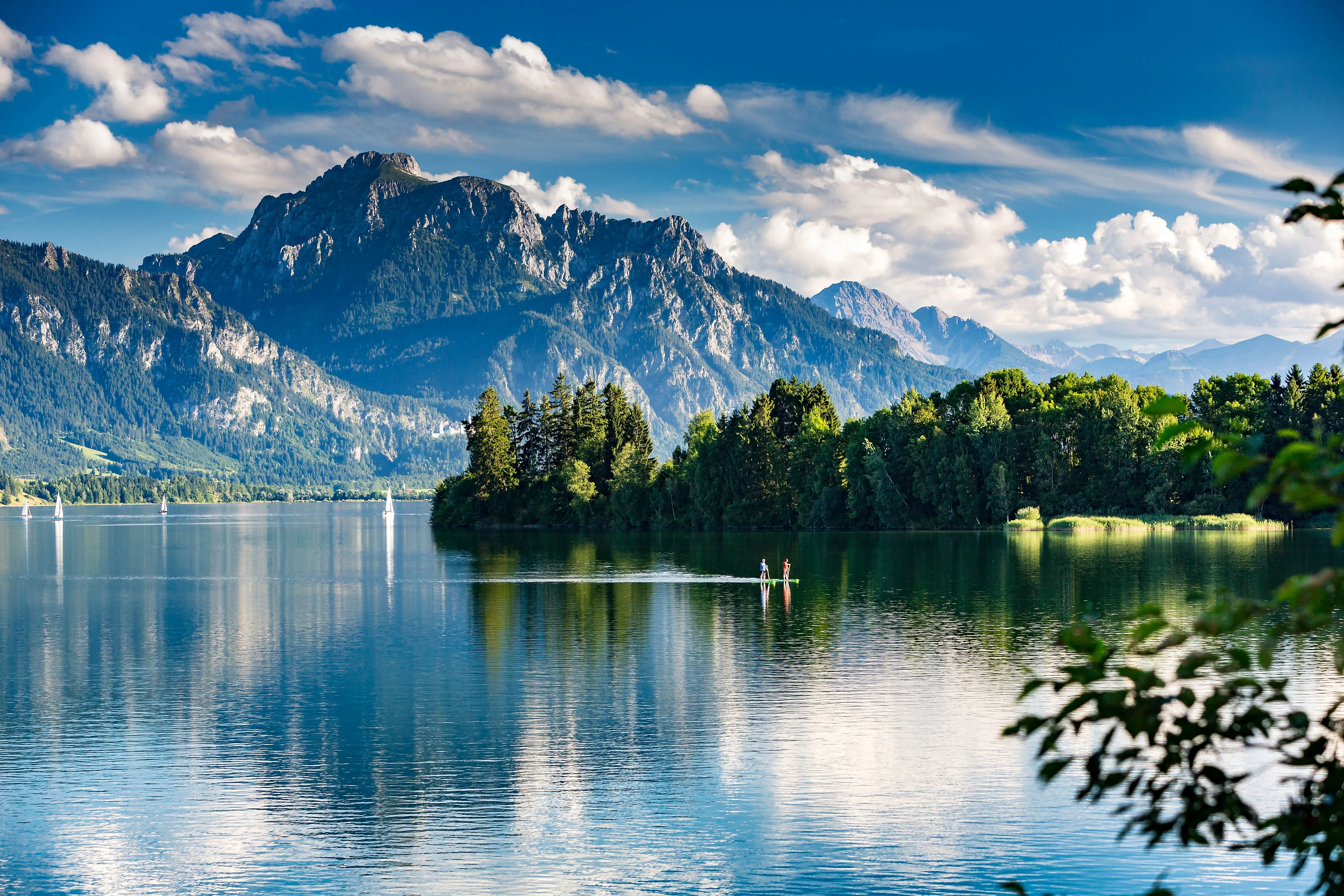 Vue sur le lac Forggensee dans l'Allgäu, avec des voiliers sur l'eau, une végétation luxuriante sur les rives et un imposant panorama alpin en arrière-plan.
