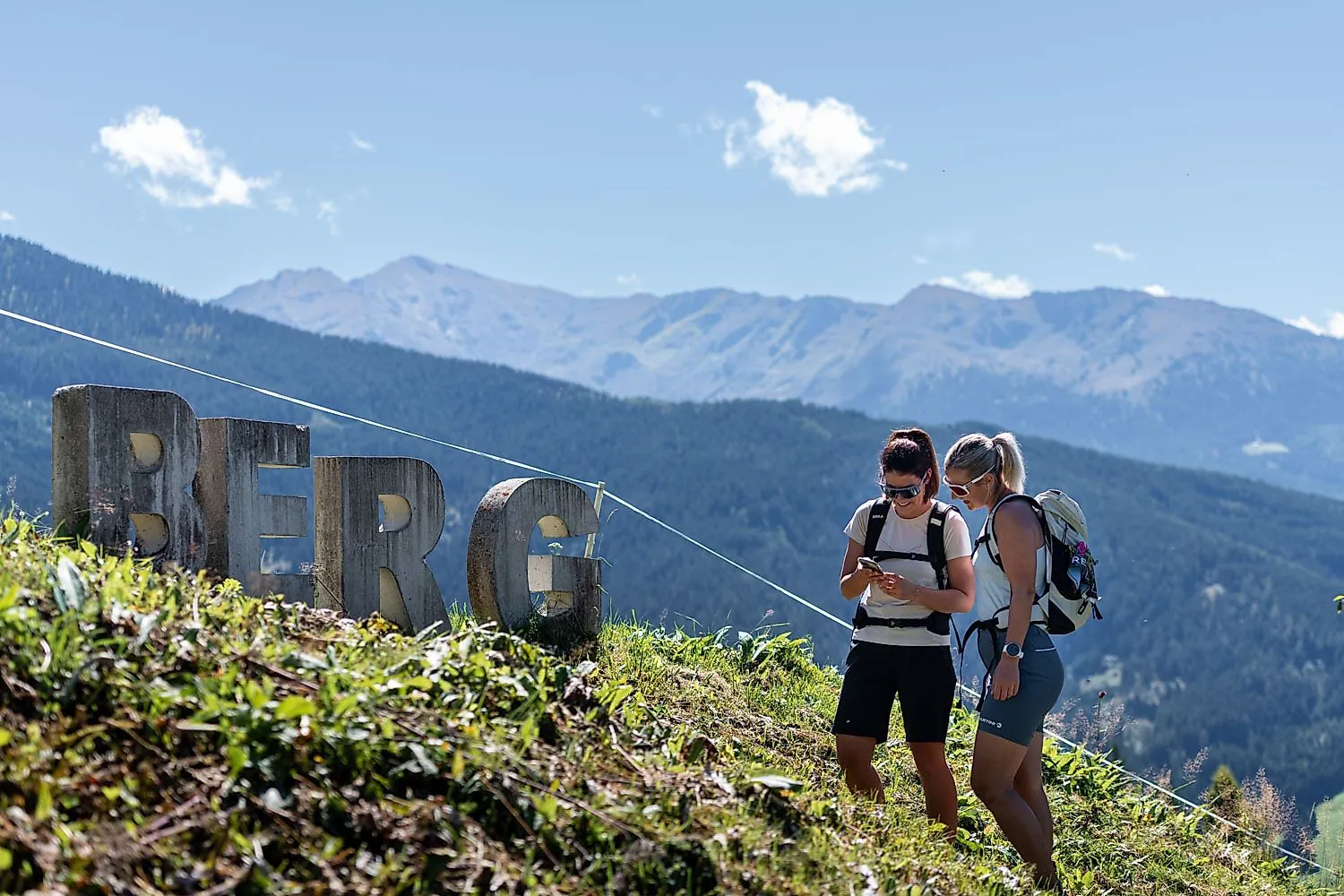 Dos excursionistas en la montaña con una vista de los Alpes y las letras de la montaña en primer plano con tiempo soleado