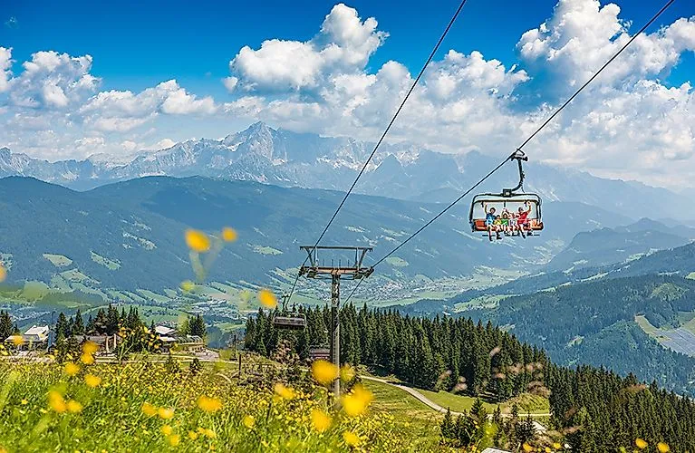 Letná horská krajina vo Flachau s rozkvitnutými lúkami, lanovkou a výhľadom na Alpy v pozadí.