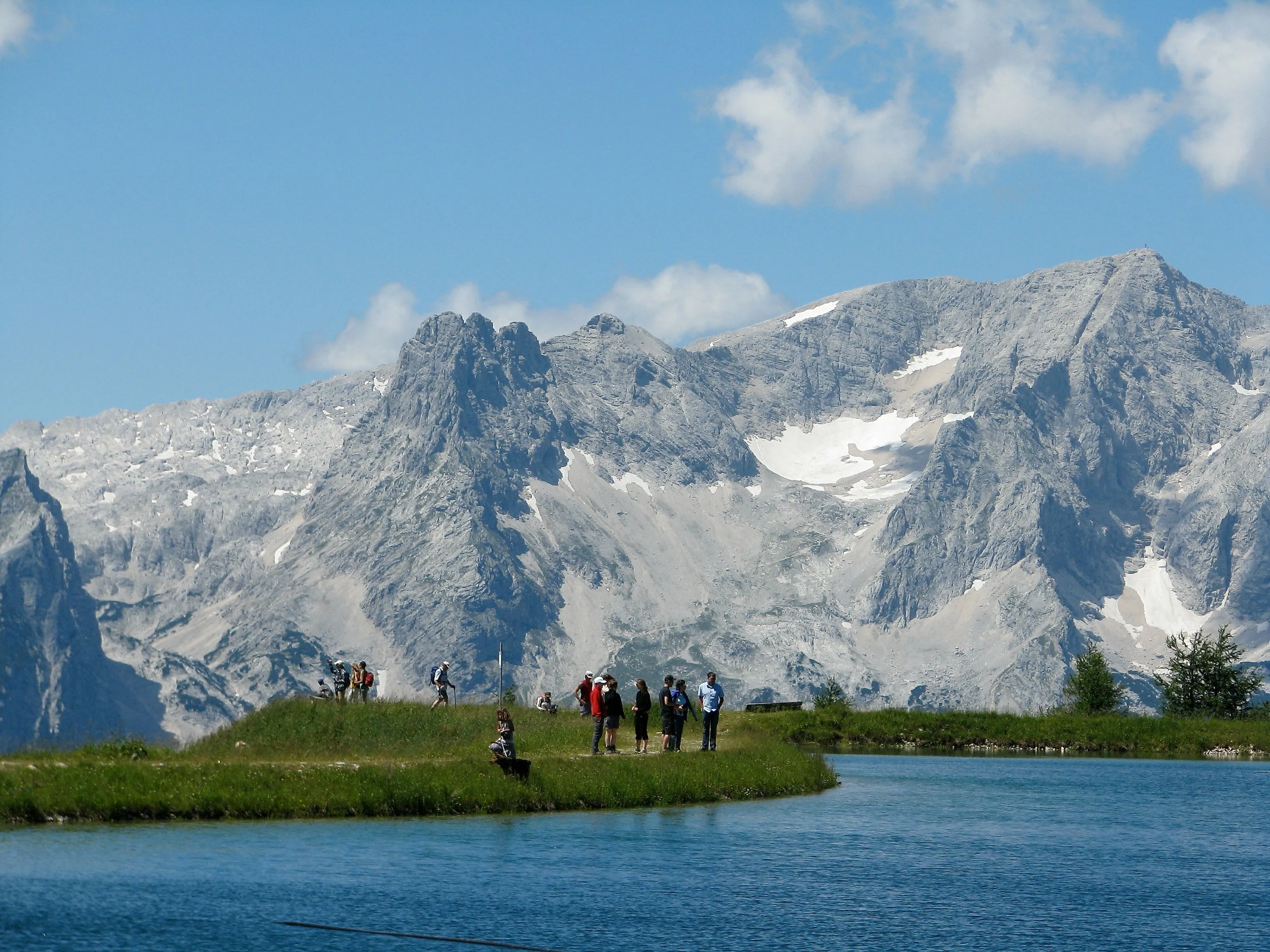Hikers against an impressive mountain backdrop on the banks of an idyllic mountain lake in Hinterstoder in clear summer weather.