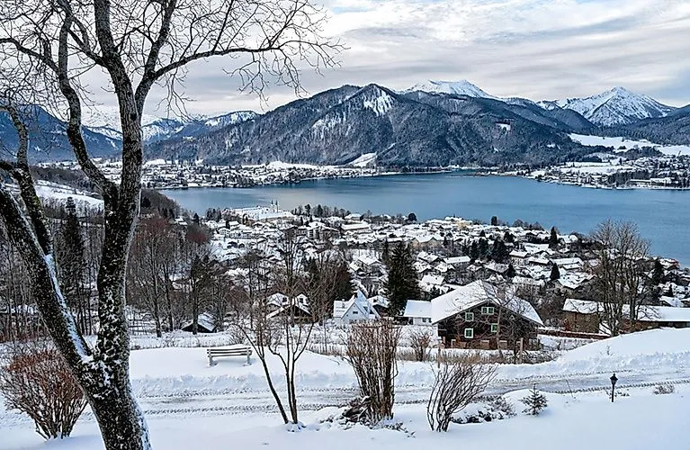 Winter panoramic shot of Lake Tegernsee with snow-covered houses and mountains in the background.