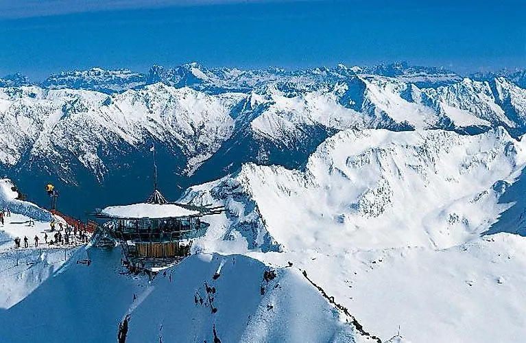 Lyžaři na vyhlídce na vrcholu Hochgurgl s působivou zimní krajinou v pozadí.