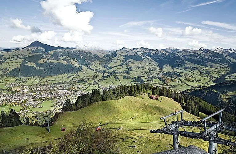 Rozsáhlý pohled na zelené Kitzbühelské Alpy s loukami, malými lesy a roztroušenými vesnicemi; v popředí lanovka s červenými gondolami.