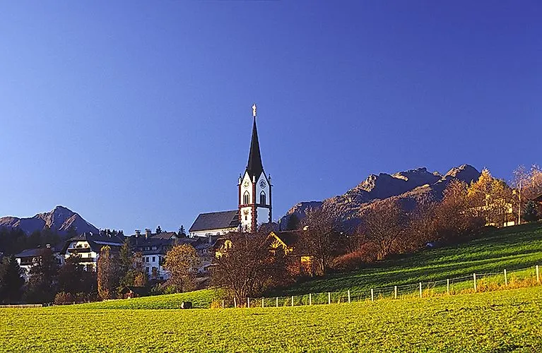 Letní panoramatický záběr Mariapfarru s pozoruhodným kostelem v popředí, zelenými loukami a pohořím Lungau v pozadí.