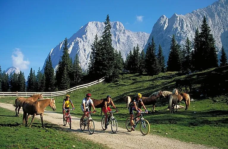 Čtyři cyklisté projíždějí alpskou loukou nedaleko Ehrwaldu, obklopeni koňmi a impozantními horami oblasti Zugspitze.